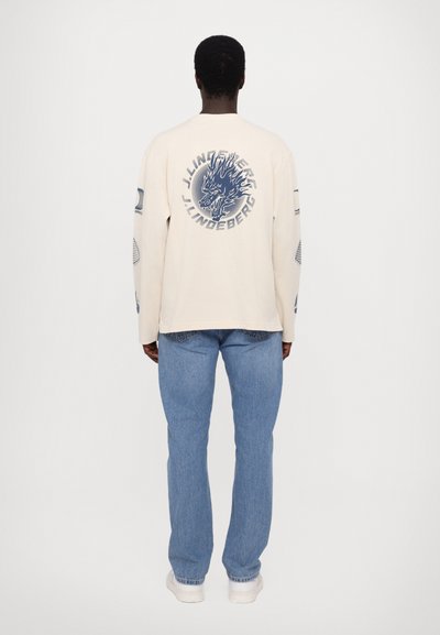 Cream long-sleeve shirt featuring a blue graphic on the back, paired with light blue jeans and white sneakers. Smooth texture throughout.