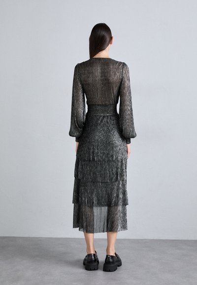 Femme avec de longs cheveux noirs portant une robe midi transparente à texture métallique et des chaussures noires épaisses, se tenant devant un mur gris uni.