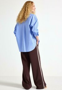 Camicia a righe azzurro chiaro con maniche a palloncino, abbinata a pantaloni marroni larghi con strisce bianche ai lati. Texture lisce visibili.