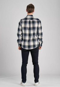 Mężczyzna stoi tyłem do kamery, ubrany w koszulę w kratę i ciemne dżinsy, na neutralnym tle.