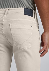 Blugi din denim bej cu o textură netedă, având un buzunar posterior cu un model de cusătură în formă de chevron și o etichetă întunecată la talie.