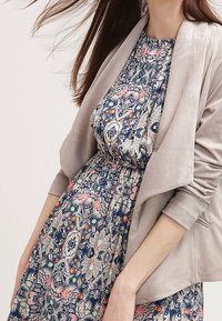 Kvinna i en blommönstrad klänning med samlad midja och en ljusbeige öppen jacka med uppvikta ärmar.