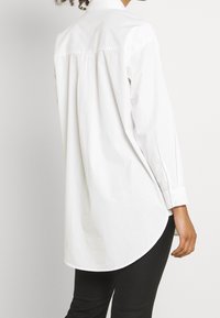 Bílá dlouhá košile s rukávy vyrobená z bavlny, s kulatým lemem, vybavená strukturovaným límcem a viditelnými manžetami s knoflíky na zápěstích.