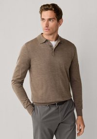 Bruine polo met lange mouwen en een gebreide textuur, voorzien van een knoopsluiting met drie knopen, gecombineerd met grijze pantalons en een zwarte riem.