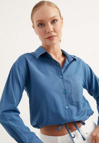 Camicia azzurro chiaro con colletto, dotata di una tasca frontale e lacci in vita. Realizzata in un tessuto liscio con maniche lunghe.