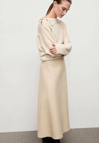 Suéter de lana beige con cuello vuelto y detalle de botones en el lateral, combinado con una falda midi de línea A a juego, ambos con una textura suave.