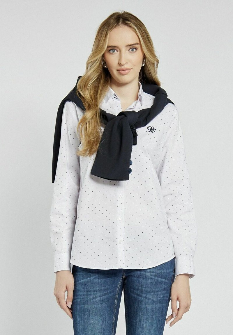 Biała koszula z guzikami, w granatowe kropki, z haftowanym logo, w połączeniu z ciemnoniebieskim swetrem zarzuconym na ramionach. Długie rękawy.