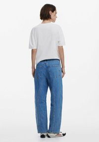 White short-sleeve t-shirt paired with blue denim jeans, featuring a relaxed fit, front pockets, and unique seam detailing. Casual footwear visible.