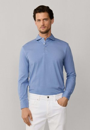 Man draagt een lichtblauw poloshirt met lange mouwen en witte broek, met één hand in de zak, staand tegen een effen grijze achtergrond.