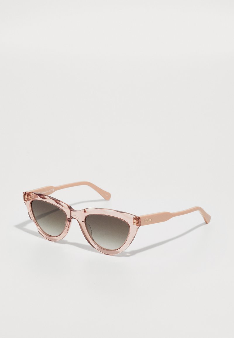Lunettes de soleil en forme d'yeux de chat avec une monture translucide rose et des verres dégradés gris foncé. Elles disposent de branches légères et d'un accent de logo subtil sur le côté.