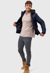 Marineblå pufferjakke med hætte, grå t-shirt, grå jeans med knædetaljer og brune støvler. Modellen poser med jakken åben og smiler.