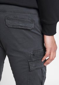 Cargo kalhoty v tmavě šedé látce mají boční kapsy, kapsu na zip a texturované švy podél švů.