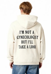 Felpa beige con una grande stampa sul retro in testo nero che recita: "NON SONO UN GINECOLOGO MA DARÒ UN'OCCHIATA." Materiale morbido, vestibilità casual.