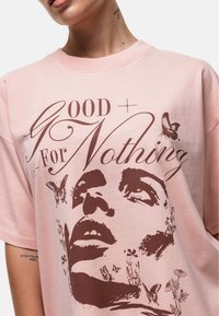 Jasnoróżowa, oversize T-shirt z bawełny, z ciemnoczerwonym nadrukiem przedstawiającym twarz, motyle i kwiatowe wzory. Okrągły dekolt.