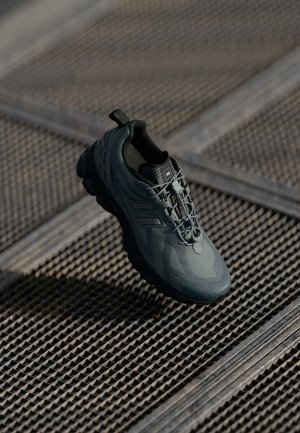 Ciemnoszary but sportowy z sznurowadłami, zawieszony nad teksturowanym metalowym rusztem w tle, podkreślający jego design i szczegóły podeszwy.