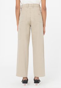 Beige, vida benbyxor i bomull med hög midja, som har bakfickor och en sidokontaktficka. Bärs med svarta sneakers.