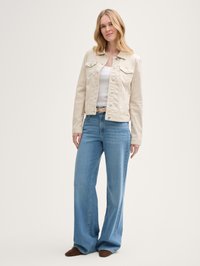Woman wearing a beige buttoned jacket, white tank top, light blue high-waisted wide-leg jeans, beige belt, and brown shoes standing against white background.
