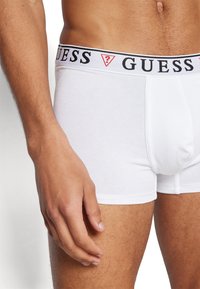 Weiße Baumwoll-Boxershorts mit einem schwarz-weißen "GUESS"-Logo-Bund. Nahaufnahme mit einer Hand, die auf dem Oberschenkel ruht.