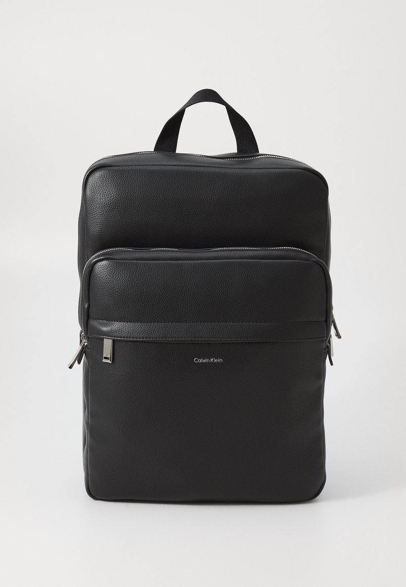 Mochila de cuero negro con una superficie texturizada, que cuenta con dos compartimentos con cremallera y un asa en la parte superior. Sutil logo de Calvin Klein en la parte frontal.