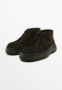 Braune Wildleder-Stiefeletten mit glatter Oberfläche, ausgestattet mit dicken grünen Gummisohlen und klassischem Schnürdesign. Nahtdetails entlang der Kanten.