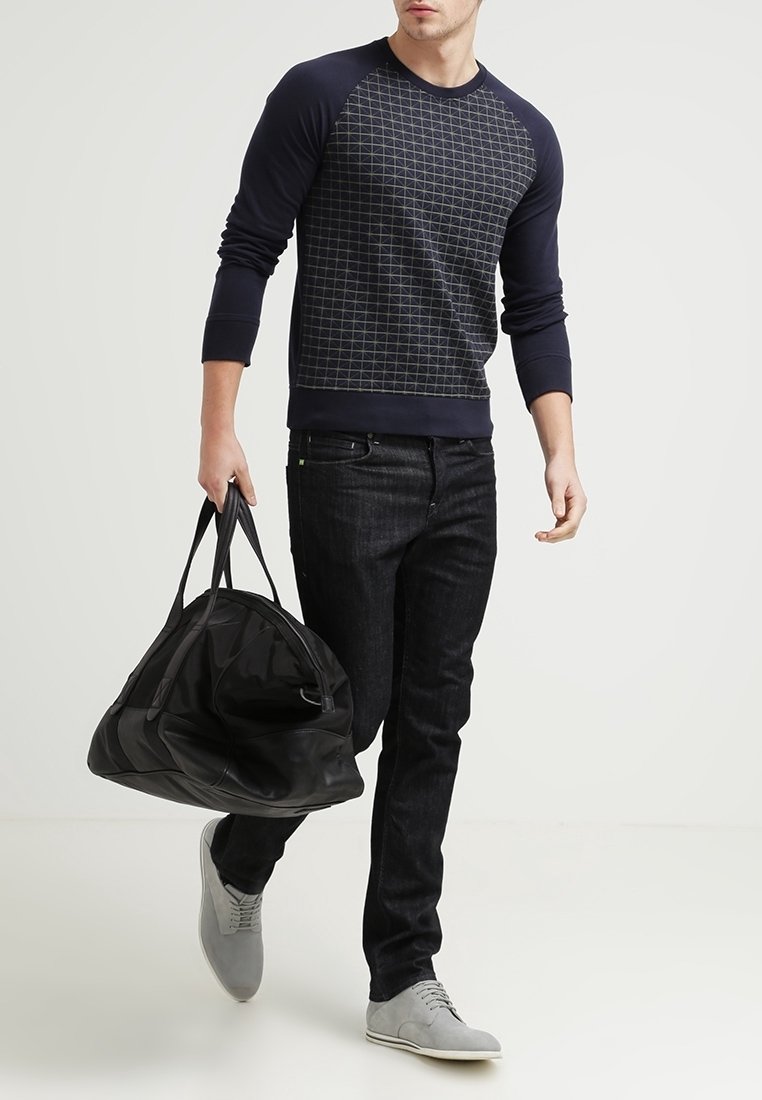 Hombre caminando con un suéter de manga larga azul marino con estampado, pantalones vaqueros negros, zapatos grises, y llevando una gran bolsa de viaje negra con la mano izquierda.