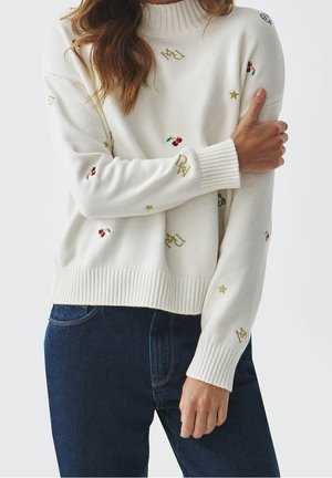Femme portant un pull blanc à col roulé avec de petites cerises, étoiles et lettres brodées, associé à un jean bleu foncé.