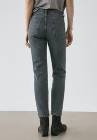 Person som bär tajta grå jeans och mörka ankleboots, stående med ryggen mot en enkel ljus bakgrund.