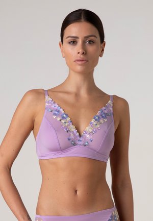 Donna che indossa un reggiseno lavanda con ricami floreali blu e gialli, in piedi davanti a uno sfondo grigio semplice.