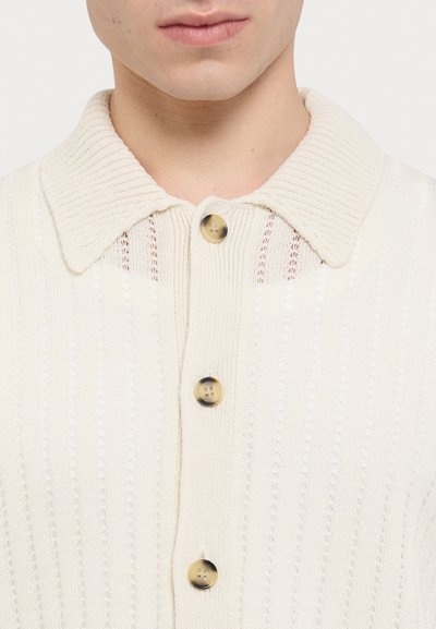 Cream-colored knitted cardigan with a ribbed collar, featuring horizontal stripes, three buttons, and a textured pattern.