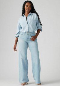 Vaaleansininen denim-paita napituksella, yhdistettynä korkeavyötäröisiin vaaleansinisiin leveisiin farkkuihin. Suuntainen pinta, rento tyyli.