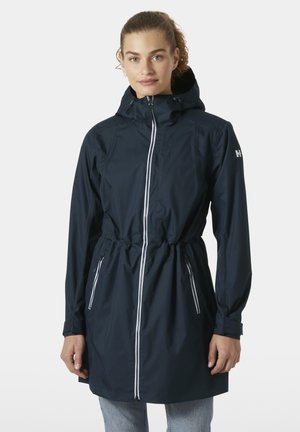 Frau trägt eine marineblaue wasserdichte Kapuzenjacke mit weißen Reißverschlüssen und Kordelzug in der Taille, stehend vor einem schlichten hellen Hintergrund.