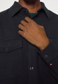 La mano di un uomo che regola il colletto di una giacca di camicia di un blu scuro con taschini sul petto e polsini con bottone, dettaglio di una piccola bandiera sulla manica.