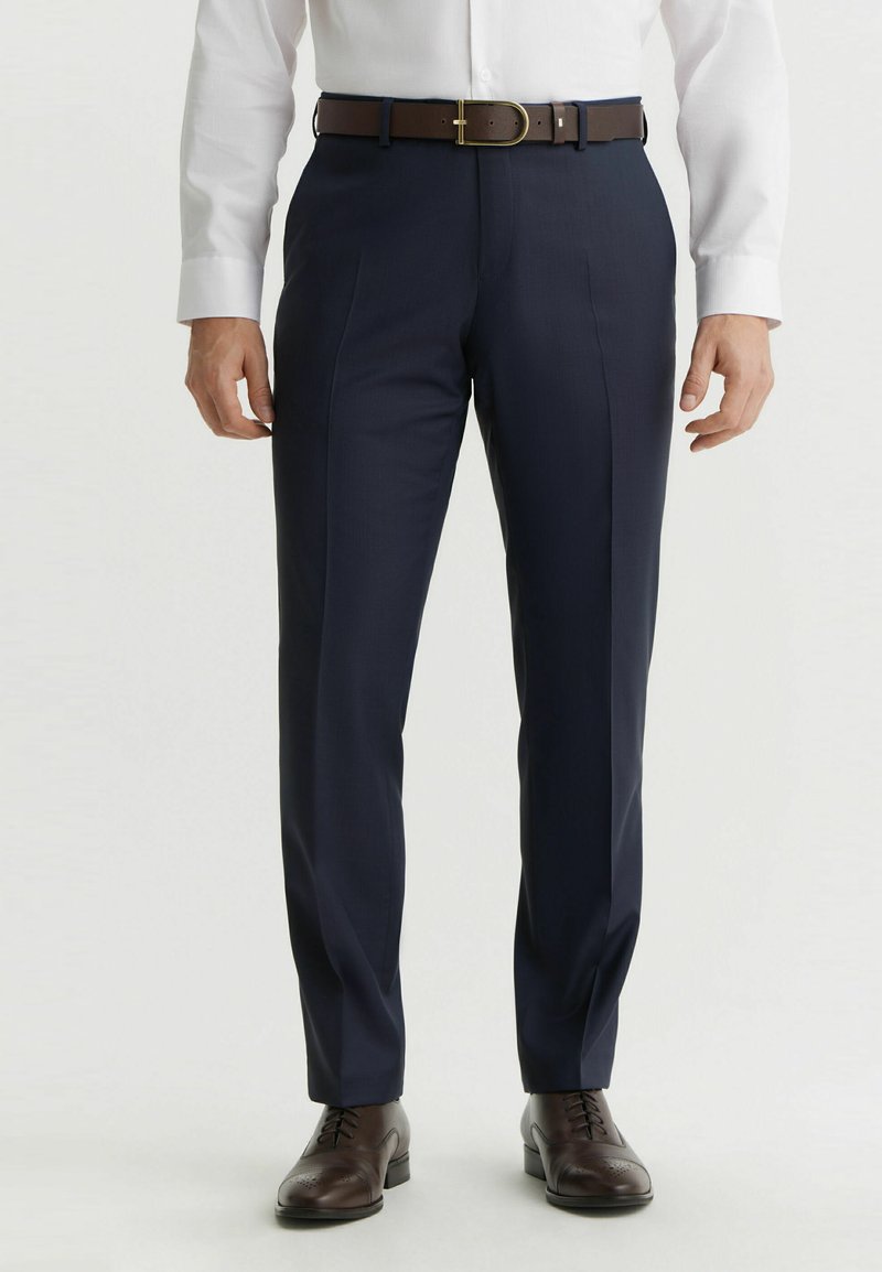 Granatowe spodnie wizytowe o dopasowanym kroju, z drobną fakturą i przodu z zakładkami. Połączone z brązowym skórzanym paskiem i brązowymi butami z detalami typu wingtip.