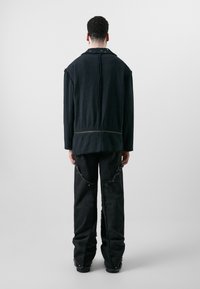 Schwarze Jeansjacke mit ausgefransten Kanten und Druckknopfdetails, kombiniert mit lockeren schwarzen Hosen mit abgenutzten Akzenten und sichtbaren Nähten.