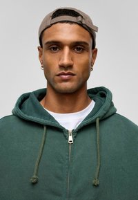 Groene hoodie met rits en verstelbare koorden, gemaakt van zachte stof. G gedragen over een wit shirt, gecombineerd met een lichtgrijze pet.