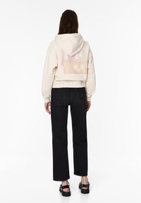 Krémová krátká mikina se vzorovaným nápisem na zádech, kombinovaná s černými džíny s vysokým pasem a černými sandály.