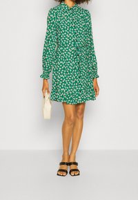 Groene bloemenjurk met knopen aan de voorkant, lange mouwen en een strikband in de taille. Bevat witte en gele bloemen op een groene achtergrond. Accessoires: crèmekleurige tas, zwarte sandalen.