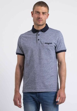 Homme portant un polo gris à manches courtes avec un petit motif, col et poignets foncés, et un jean bleu, debout devant un fond uni.