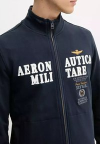 Giacca zip-up navy in misto cotone. Caratterizzata da testo stampato bianco, ricami dorati e tasche laterali. Design con colletto alto.