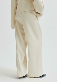 Beige vida byxor med bakfickor, bärs med en matchande beige kavaj och svarta spetsiga stilettklackar mot en vit bakgrund.