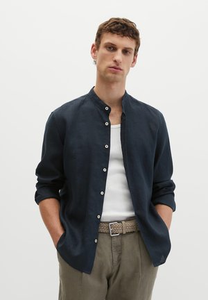 Młody mężczyzna w granatowej koszuli zapinanej na guziki, na białym tank topie, z beżowym plecionym paskiem i oliwkowozielonymi spodniami, stoi z rękami w kieszeniach.