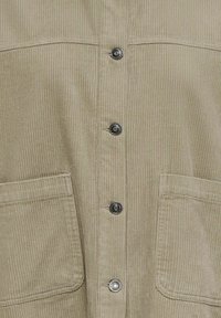 Bēša kordurojas jaka ar vertikālu ribotā raksta tekstūru, četriem metāla spiedpogu aizdares elementiem un divām lielām priekšējām līplomām kabatām.