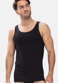 Mann in einem enganliegenden schwarzen Tanktop, stehend mit neutralem Gesichtsausdruck, leicht nach links schauend vor einem schlichten hellen Hintergrund.