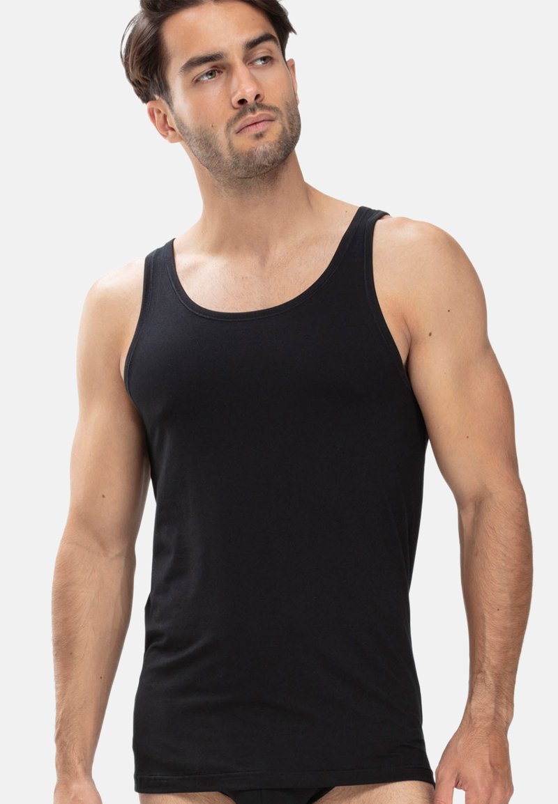 Mann in einem enganliegenden schwarzen Tanktop, stehend mit neutralem Gesichtsausdruck, leicht nach links schauend vor einem schlichten hellen Hintergrund.