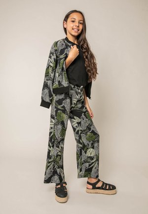 Meisje met lang haar draagt een bijpassend zwart jasje en broek met witte luipaard- en groene bladprints, een zwart topje en zwarte platformsandalen.