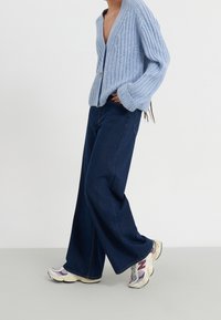 Světle modrý žebrovaný cardigan s hlubokým výstřihem do V, pře oversized rukávy, spárovaný s širokými džínami z tmavého denimu a vícebarevnými teniskami New Balance.