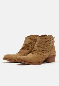 Bottines en daim beige avec un dessus perforé, un bout pointu et un talon en bois empilé. Un motif ondulé distinct dans les perforations ajoute de la texture.
