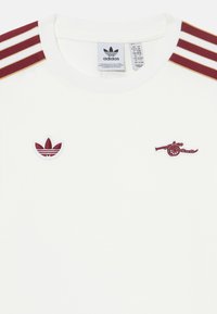 Hvit kortermet skjorte med maroon og gullstripete skuldre. Har brodert Adidas-logo og kanoninsignia på brystet.