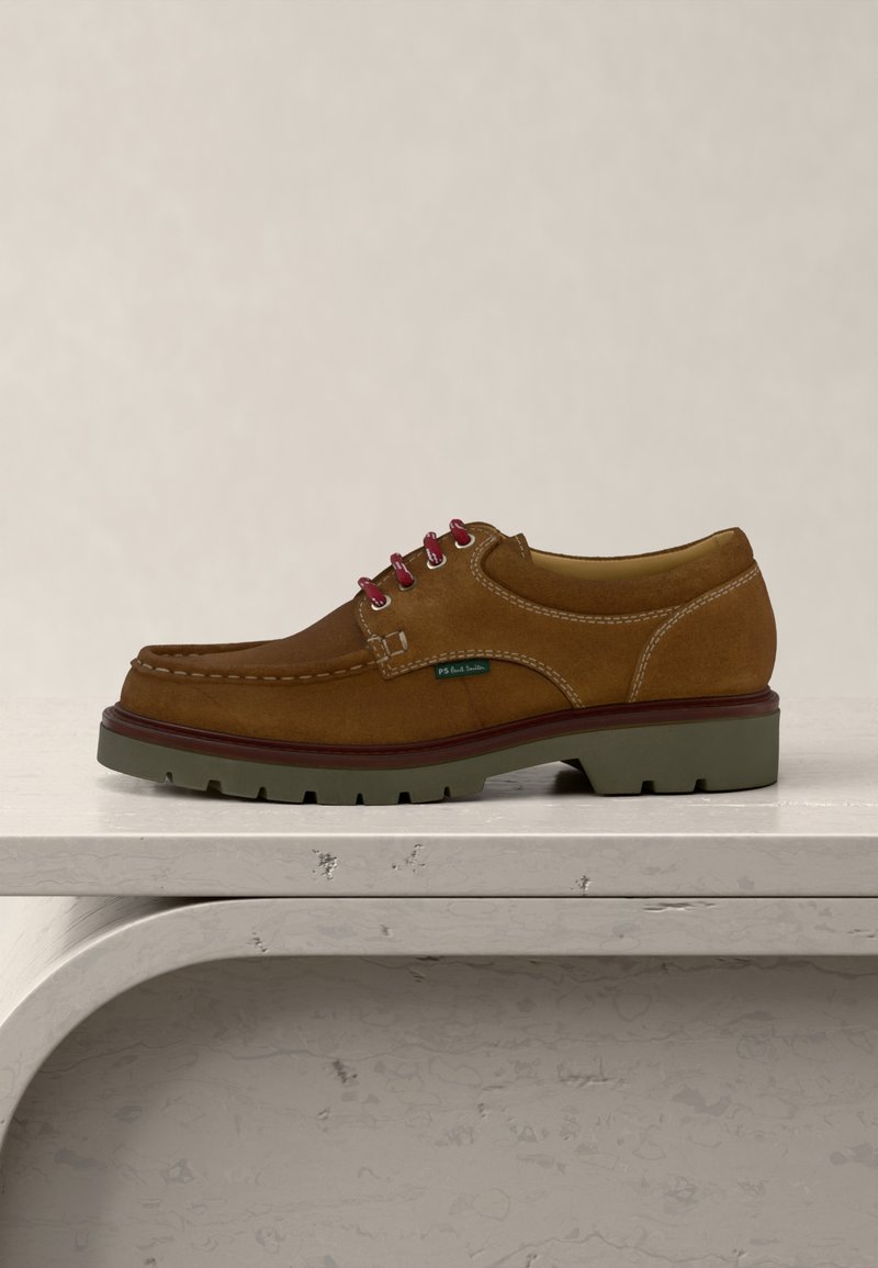 Chaussure en suède marron à lacets avec œillets rouges, semelle épaisse verte, coutures blanches, et étiquette "PS Paul Smith" sur surface blanche.
