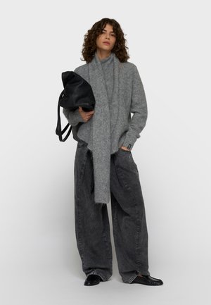 Kvinde med krøllet hår iført en grå fluffy tørklæde og sweater, brede mørkegrå bukser, sorte loafers, der holder en sort læderrygsæk.